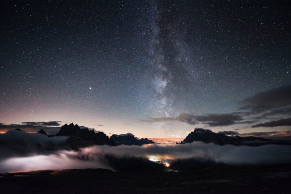 You can clear see the milky way. Something is shining in the middle of the forest with stars in the sky. The mountains are surrounded by dense fog.