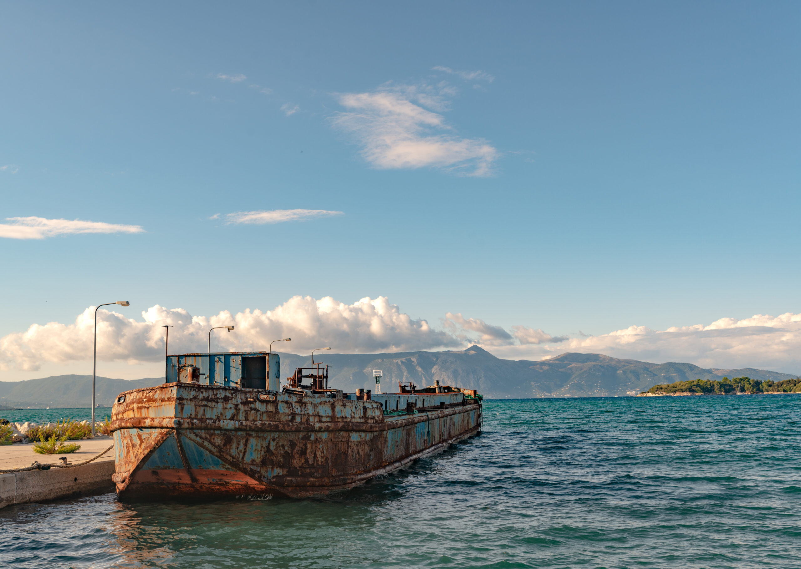 View of old rusty argosy near pier in Ionian sea. Beautiful landscape of Corfu island, mountains on background in sunny day.