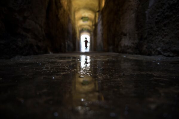 A figure stands at the end of a dark, stone tunnel. The floor is wet, and the tunnel walls show age. The light at the end of the tunnel creates a stark contrast.