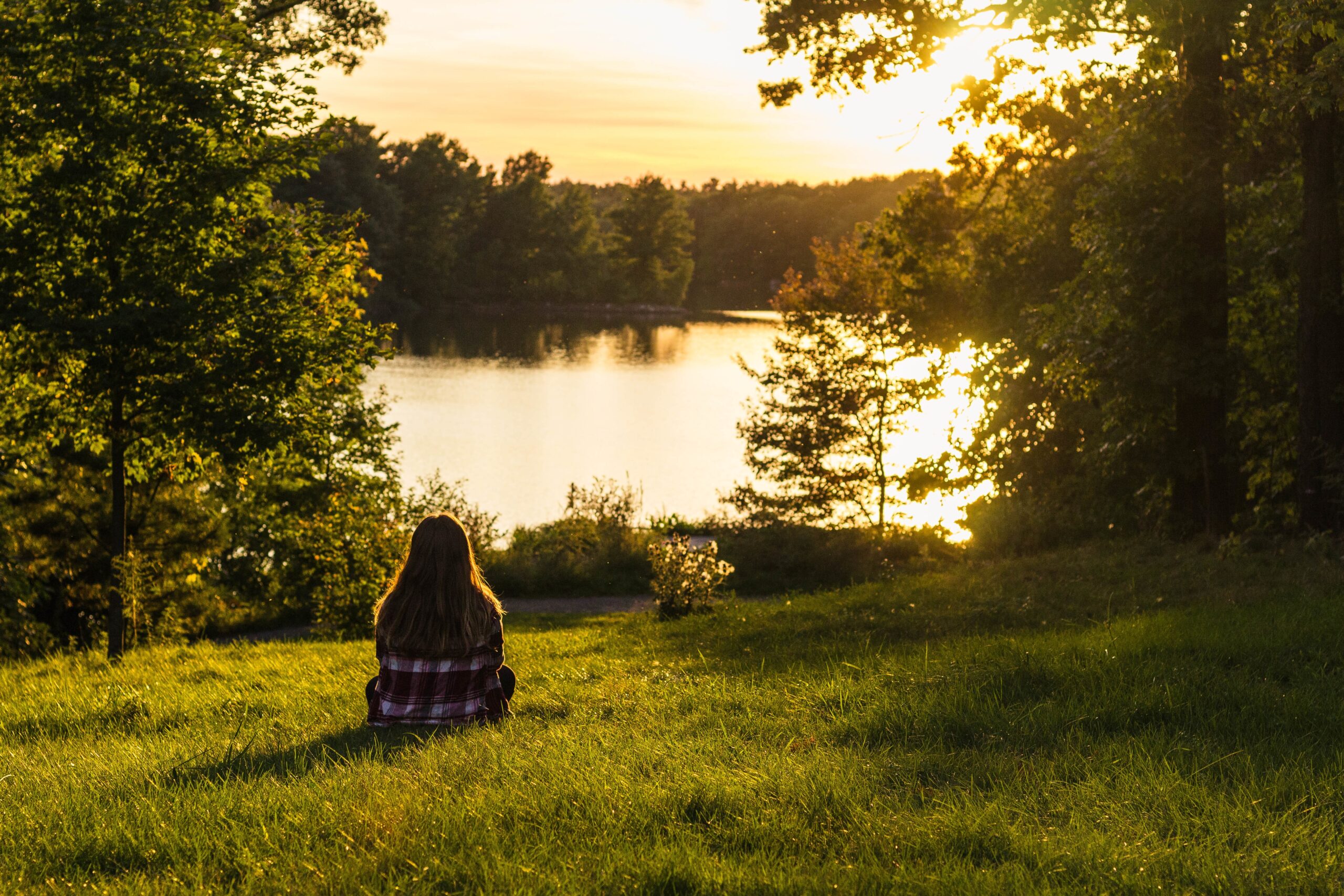A woman sitting on the grassy field enjoying the view with sea and trees in the background
